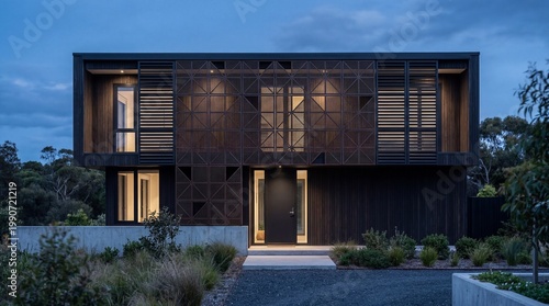 Modern house with illuminated geometric facade and dark cladding at twilight set in a natural landscape with lush vegetation.
