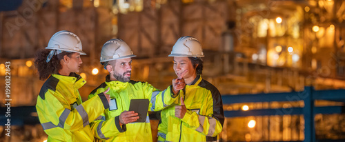 Petroleum engineers team wearing safety uniform and hardhat with laptop monitoring and discussing work at oil refinery site during night operation engineering teamwork in energy industry.