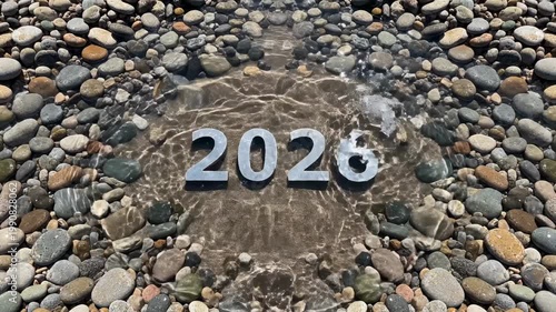 Silver numbers 2026 partially submerged in clear water surrounded by smooth pebbles on a sandy beach, creating a serene and reflective atmosphere