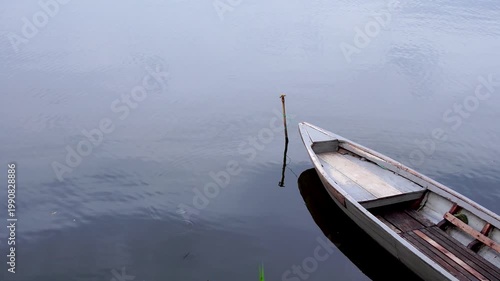 Bow of a traditional wooden fishing boat tethered to a bamboo pole in the calm waters of Selorejo Reservoir, Malang, Indonesia, depicting the quiet reality of rural Southeast Asian livelihoods.