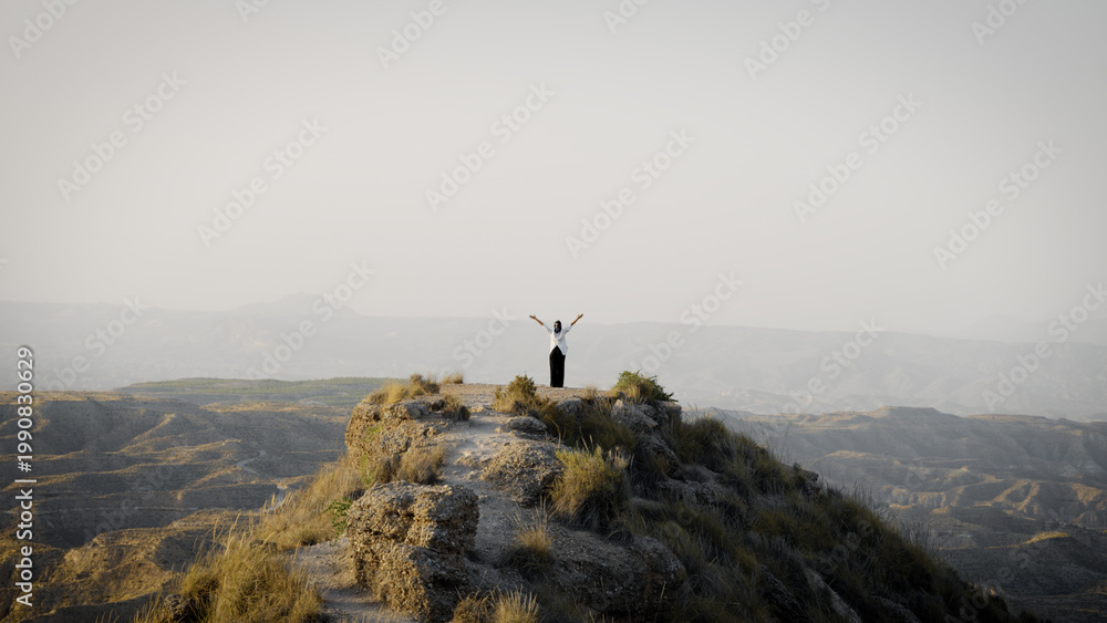 Obraz premium Muslim woman with arms outstretched on a desert mountain peak, embracing nature and feeling joy and spirituality