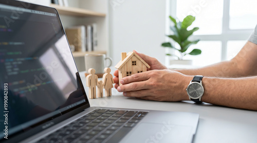 Hands Protecting Wooden House Model Next to Family Figurines and Laptop