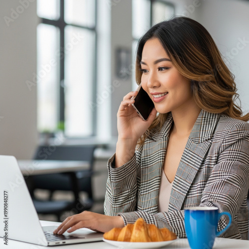 A smiling young businesswoman sits at her office desk talking on a mobile phone while working on her laptop computer