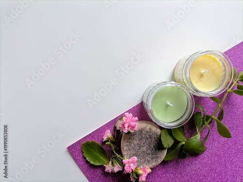 Candles glow softly beside pink blossoms. Green and yellow waxes rest on glittery purple mat. Leaves and petals add gentle, natural charm. Scene evokes calm, quiet relaxation