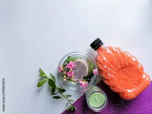Soft spa setup with glowing candle, pink flowers, and orange bath salts. Calm, soothing tones evoke relaxation and self-care. Glass bowl holds mint, petals, and lavender for aromatherapy