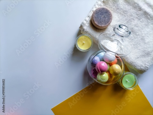 Colorful bath bombs and candles rest on soft towel. Glass jar holds pastel spheres, hinting at relaxation. Yellow and white surfaces create clean, soothing contrast. Still life evokes calm