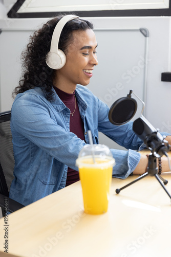 Non-binary podcaster speaking into mic in studio wearing denim and headset with orange juice