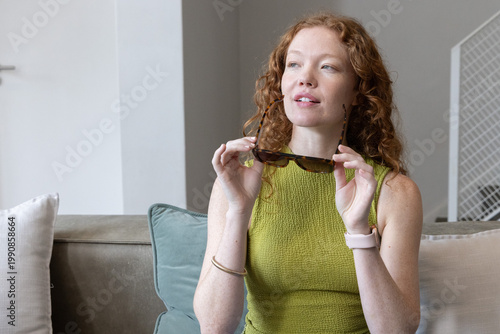 Woman sitting on sofa holding tortoise-sunglasses, wearing olive-green top showing smartwatch