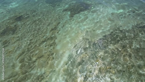 Underwater scene showing clear water over coral reef with visible sandy bottom and sunlight reflections creating patterns on the ocean floor