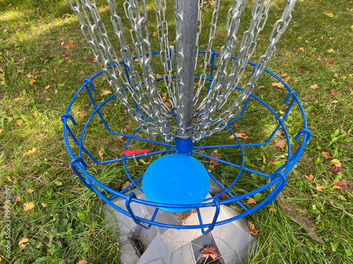 Detailed view of blue circular target basket and silver chains for disc golf. Metal equipment for outdoor sports activity located on green lawn with fallen autumn leaves.