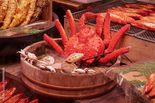 A market scene shows a large red crab placed in a wooden container. Various seafood items surround it. The setting is lively with many colors and textures