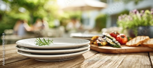 Plain ceramic plates stacked beside fresh herbs, sliced bread, and grilled vegetables on clean outdo