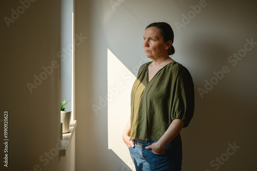 Pensive woman illuminated by soft window light, looking out the window with introspective expression, emotional lifestyle portrait in a calm home interior.