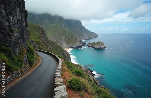 Winding road hugs cliff edge above dramatic sea coast. Green mountains meet blue ocean waters under cloudy sky. Coastal landscape with rocky shore and turquoise waves.