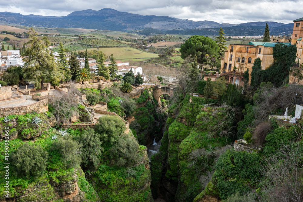 Obraz premium High angle panoramic view of the lush El Tajo gorge in Ronda with white houses and historic architecture perched on steep limestone cliffs.