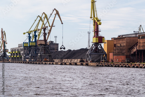 Industrial port with large cranes loading bulk materials by the water. Harbor infrastructure used for cargo handling, shipping and heavy industry operations