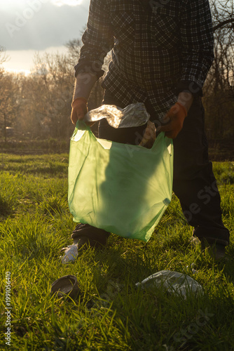 A person bends down to pick up trash and put it into a green bag. The sun shines in the background. Grass covers the ground, and trees can be seen in the distance