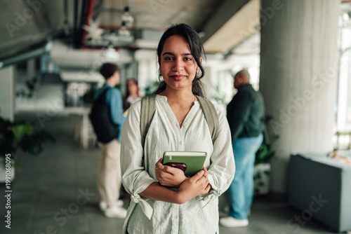Young indian woman studying, holding books, wearing backpack, standing in a university hallway, looking at camera, smiling