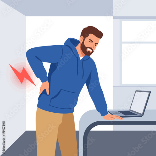 A young man in a hoodie shows discomfort due to back pain while working on his laptop at a desk, highlighting the importance of ergonomics in daily work life for wellness.