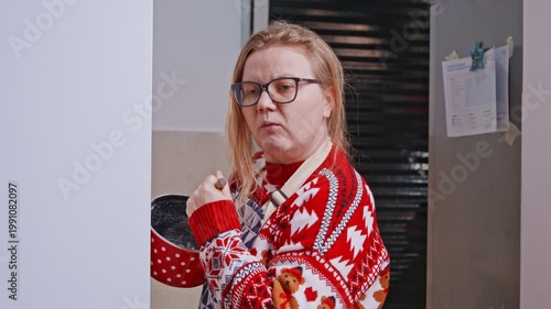 A grown woman in a red sweater with a flour stained face peers out from around the corner holding a red saucepan with a whisk in the kitchen, looking away in surprise.