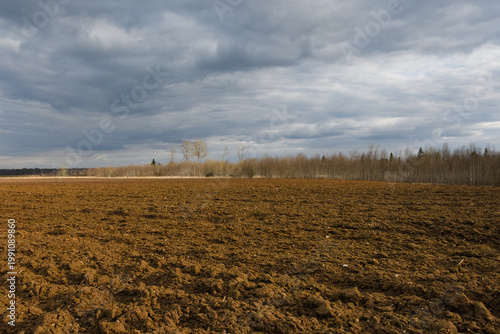 Plowed farm field with rich soil texture under a moody spring sky. Natural light gradient illuminates the brown earth and distant forest, creating a dramatic rural landscape.