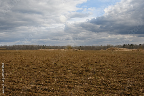 Plowed agricultural field in spring under a dramatic cloudy sky. Textured brown soil and distant piles of fieldstones near the forest line in the soft evening light.