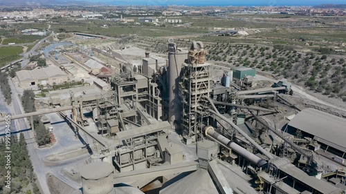 Aerial view of cement manufacturing plant in Alicante, Spain. Industrial complex with silos, conveyor systems and production facilities surrounded by agricultural landscape near Mediterranean coast. 