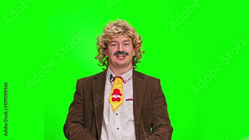 Mature man with curly blond wig and moustache wearing brown jacket and funny Christmas tie pointing at camera against green background, comic presenter concept, entertaining scene from studio life.