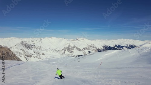 A skier in bright green jacket skis down a snowy mountain slope with snow capped mountains in the background. Slow motion, clear day with blue sky