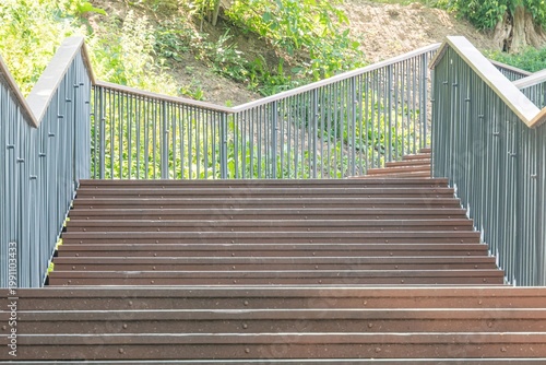 A series of metal stairs ascending a hill amidst lush greenery, with a railing on either side The perspective is from the ground looking up