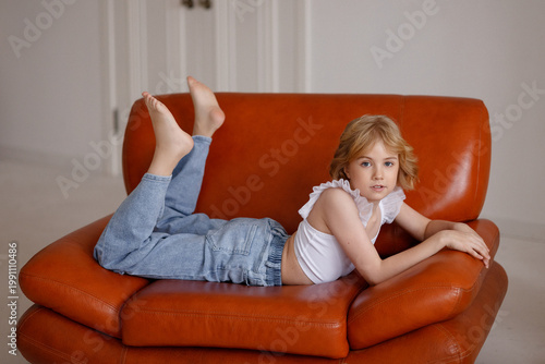 Smiling child in white sleeveless top and jeans lying on orange leather sofa in bright living room, side view, cozy home atmosphere, childhood, lifestyle, relaxation, weekend, advertising concept.