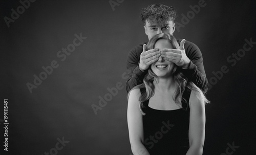 Фотография Young man standing behind happy woman, covering her eyes with hands, anticipatio