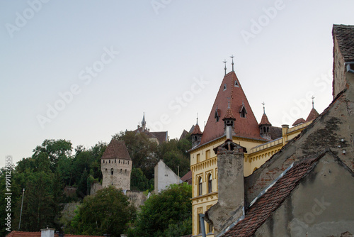  Antique castle towers rise above rustic rooftops at sunset, capturing the essence of historical European architecture. Weathered brick details and lush greenery add to the scene's charm.