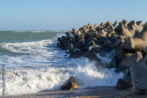 Powerful ocean waves crash against large concrete tetrapods on a sunny day. The solid structures protect the shoreline, highlighting the energy and beauty of the sea's movement.