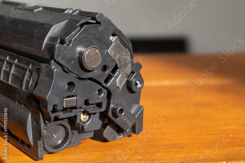 Black printer toner cartridge rests on warm wooden surface. Close-up highlights mechanical components and internal structure. Industrial design shows functional details with precision