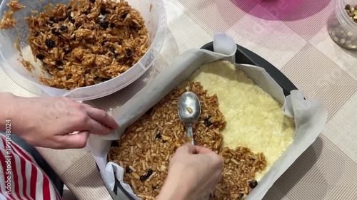 caucasian home cook smoothing and compacting granola layer over pastry base with spoon and palm, creating even bar surface, scattered oats and dark fruit visible, calm finishing step before bake