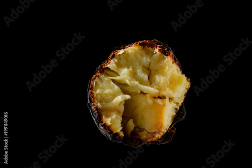 Single baked potato in skin isolated on black background. Macro view of a whole potato baked in coals split open to show fluffy yellow texture. Rustic organic food concept isolated on black