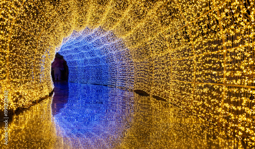 Light tunnel installation. Thousands of yellow and blue LED lights form an arched corridor. Reflections shimmer on the wet floor creating a mirrored pathway.