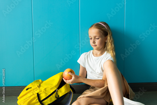 Young girl sitting on floor holding an apple next to yellow backpack