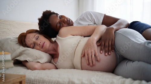 Expectant same-sex couple taking a restorative daytime nap while the non-gestational partner protectively cradles the bare baby bump.