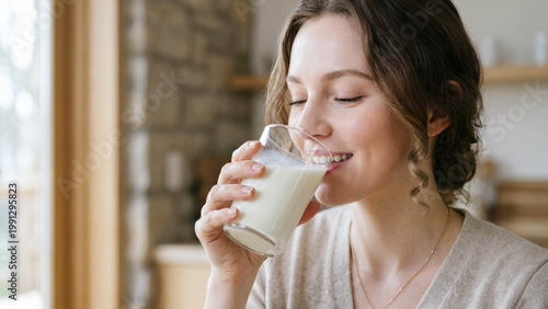 A smiling young woman with wavy hair enjoys a fresh glass of milk in a bright kitchen. The close-up shot captures her joyful expression against a soft-focus background of stone and wood textures.