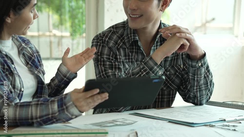 Two coworkers in plaid shirts review documents and a tablet at a desk, smiling in a bright office setting, suggesting teamwork, planning, and business collaboration.
