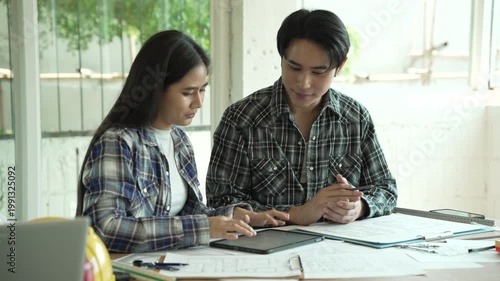Young couple collaborating at a table, reviewing documents and using a tablet in a bright home office, casual plaid shirts, planning or budgeting together