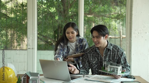 Young couple reviewing architectural plans at a bright home office table with laptop and house model, greenery visible through large windows