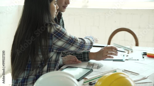 Two construction professionals in safety vests and hard hats discussing plans; female engineer holds a building model while coworker reviews clipboard at an indoor job site.