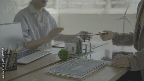 Business meeting about sustainable architecture: people discussing a miniature eco house model, ESG report on tablet, laptop, and wind turbine figurine in modern office.