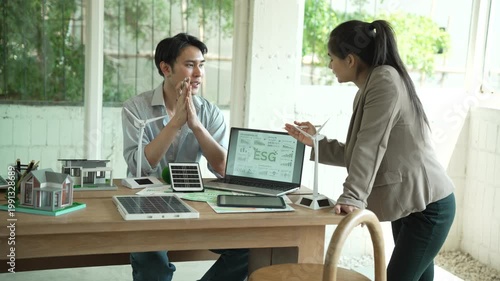 Business colleagues discuss ESG strategy at modern office desk with laptop showing ESG dashboard and renewable energy models, planning sustainable development and green finance.