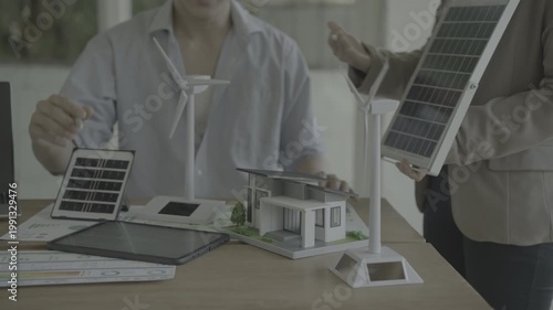 Engineers review a solar and wind energy model: miniature eco house, solar panels, wind turbines, and digital tablet on a desk in an office, showcasing renewable power planning.