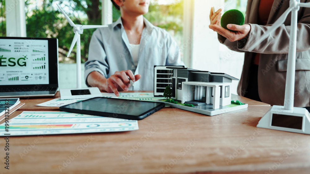 Fototapeta premium Business meeting on sustainable real estate: ESG dashboard on laptop, model eco house on desk, wind turbines and green concept props in modern office.