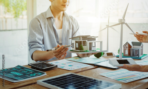 Team discussing sustainable housing model with wind turbine, solar panels, and energy charts on desk in bright modern office; renewable energy planning and green architecture concept.